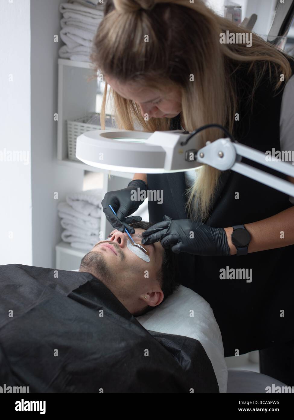 Il tecnico di bellezza sta applicando con attenzione le estensioni delle ciglia ai clienti maschi in un moderno salone, con illuminazione luminosa e spazio di lavoro organizzato che migliora la qualità Foto Stock