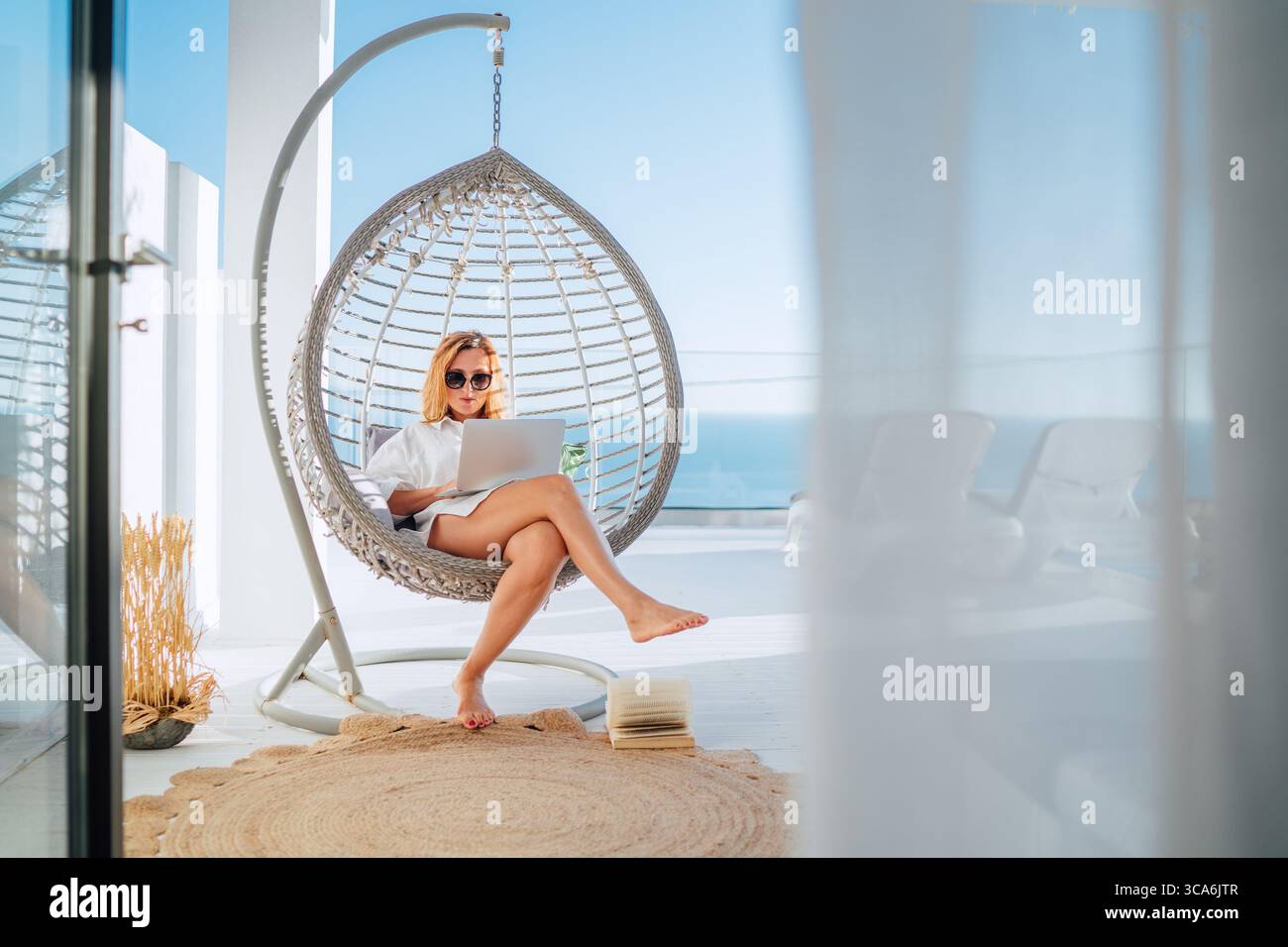 Bella donna di mezza età che si rilassa in un'accogliente sedia appesa sulla terrazza all'aperto, utilizza un computer portatile moderno, naviga su Internet e fai shopping online. Moderna Foto Stock