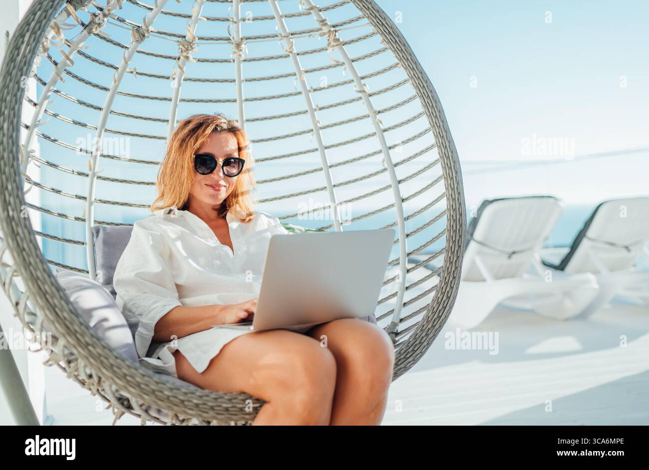 Bella donna di mezza età che si rilassa in un'accogliente sedia appesa sulla terrazza all'aperto, utilizza un computer portatile moderno, naviga su Internet e fai shopping online. Moderna Foto Stock