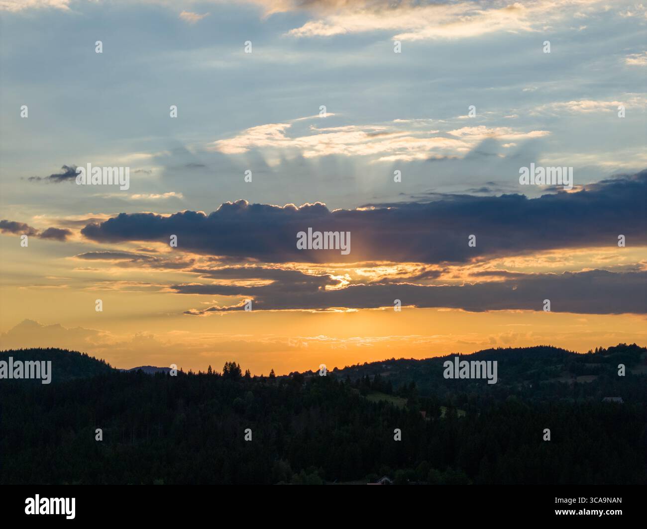 La calda luce del tramonto si irrompe attraverso nuvole a strati, diffondendo toni dorati su colline e valli boscose lontane Foto Stock