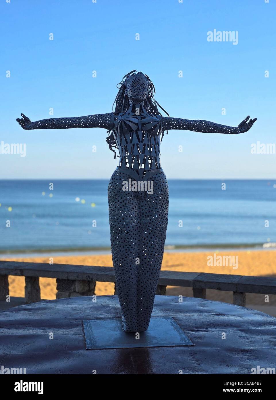 Scultura in metallo di una donna con braccia allungate che si affaccia sulla spiaggia di Albufeira in Algarve, Portogallo, con l'Oceano Atlantico sullo sfondo. - Immagine stock catturata con smartphone