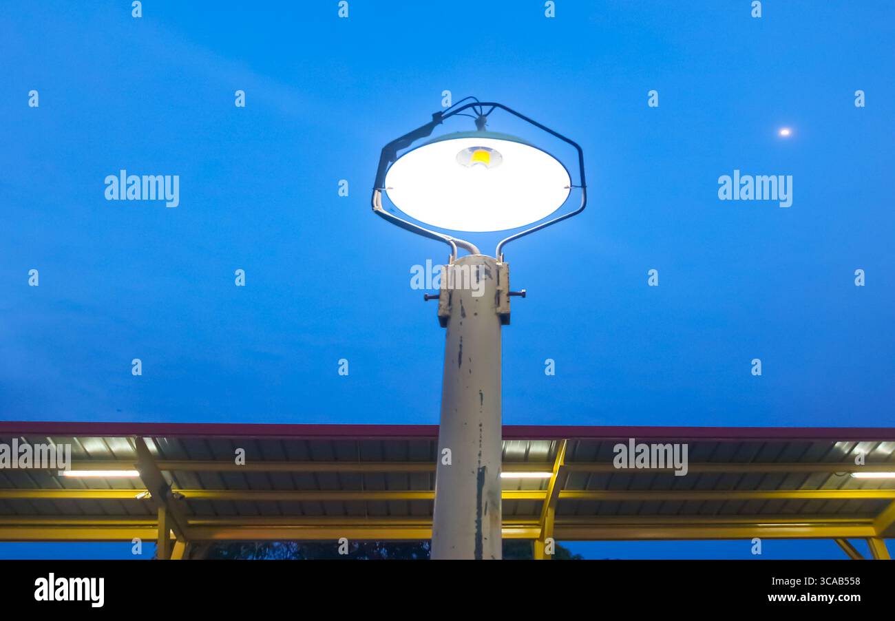 Classica lampada da strada in ferro che si illumina di bianco contro un cielo blu intenso al crepuscolo con una piccola mezzaluna, sopra le strutture del tetto gialle rosse, evocando serate tranquille Foto Stock