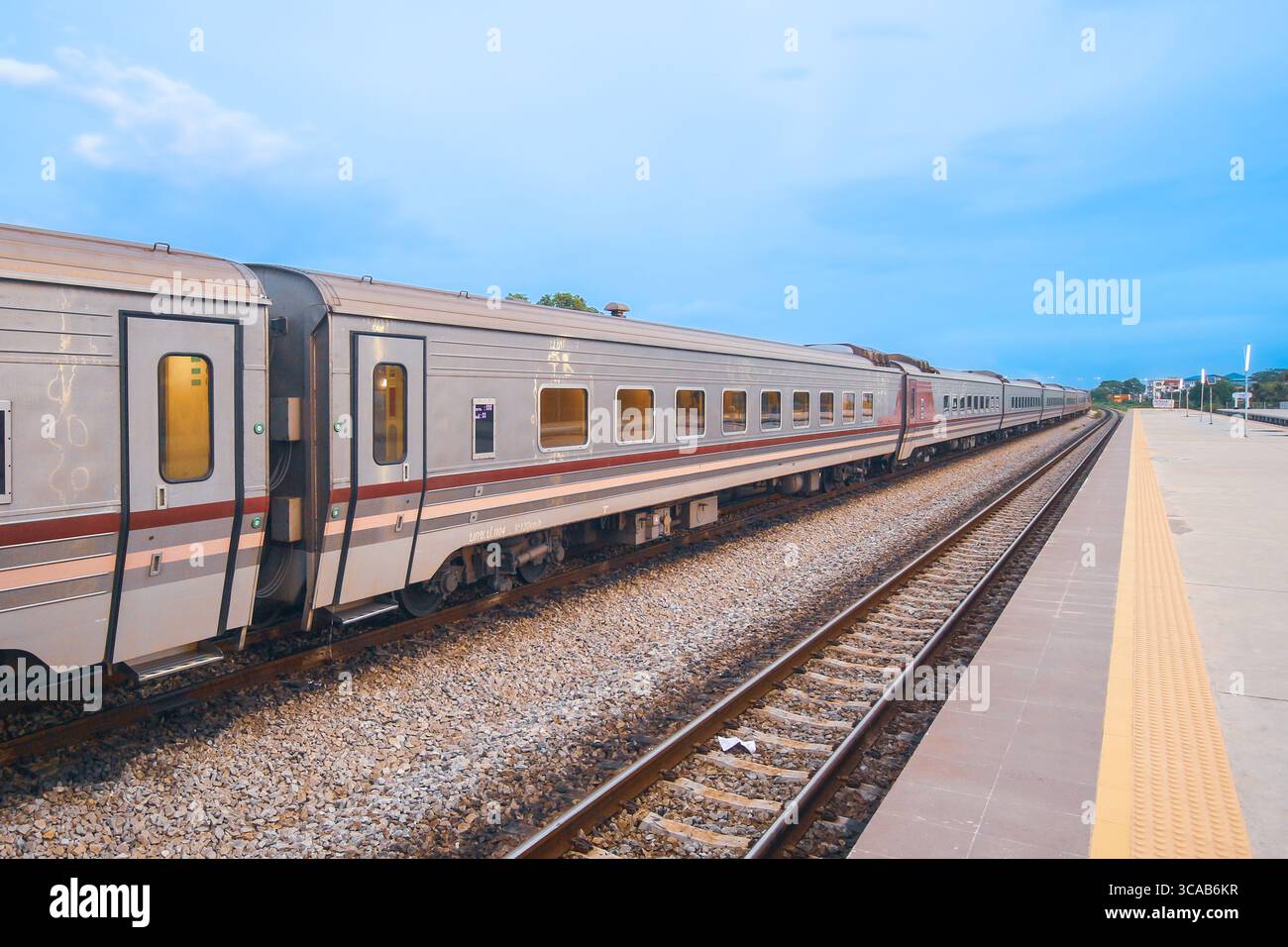 Treno passeggeri su binari curvi che conducono a una piccola città, con cielo azzurro, ghiaia e paesaggi lontani che evocano un viaggio panoramico. Foto Stock