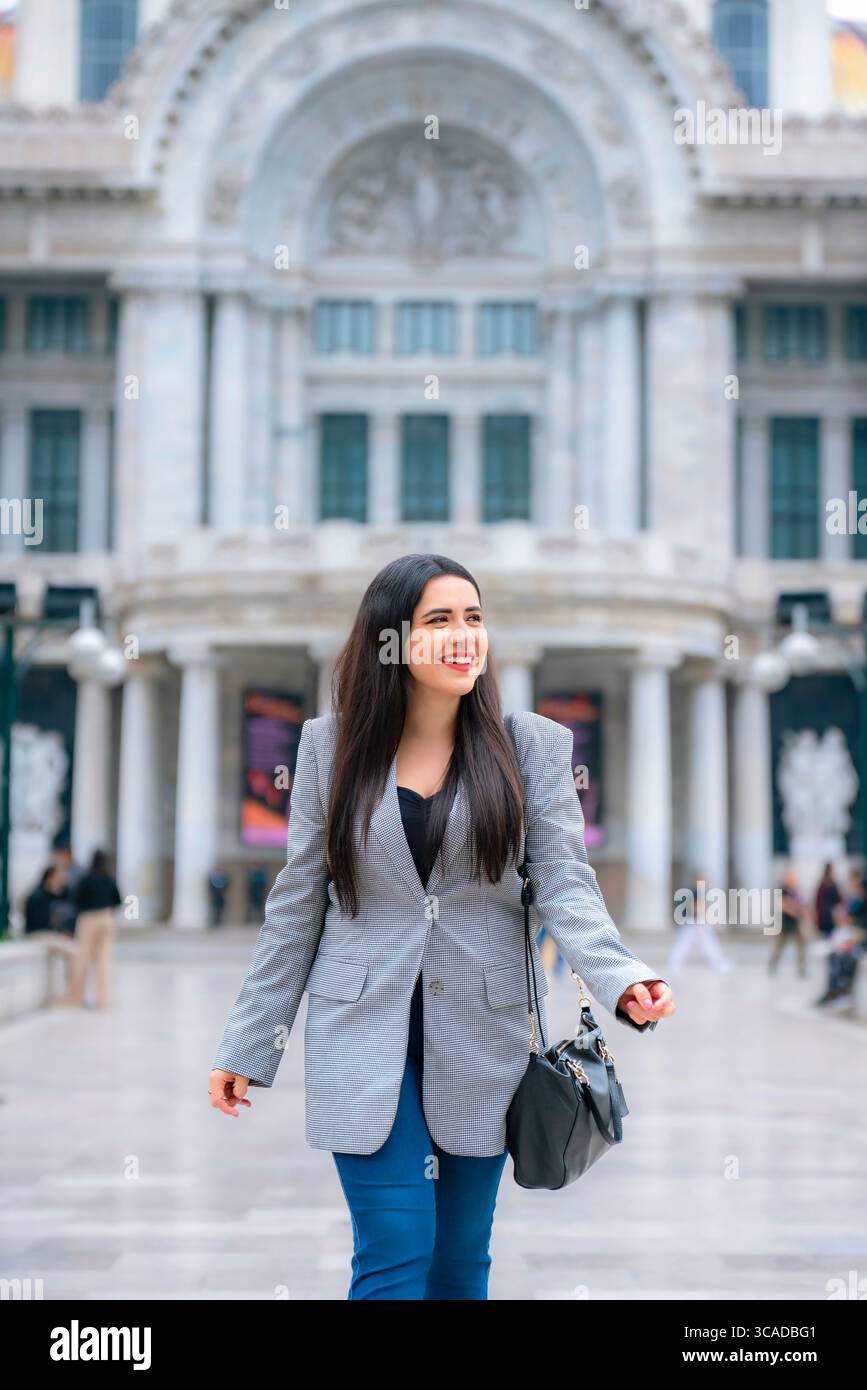 Donna latina, giovane e felice, con capelli lunghi e dritti, blazer grigio, top scuro e pantaloni blu, sorridente mentre cammina a città del Messico vicino a me Foto Stock