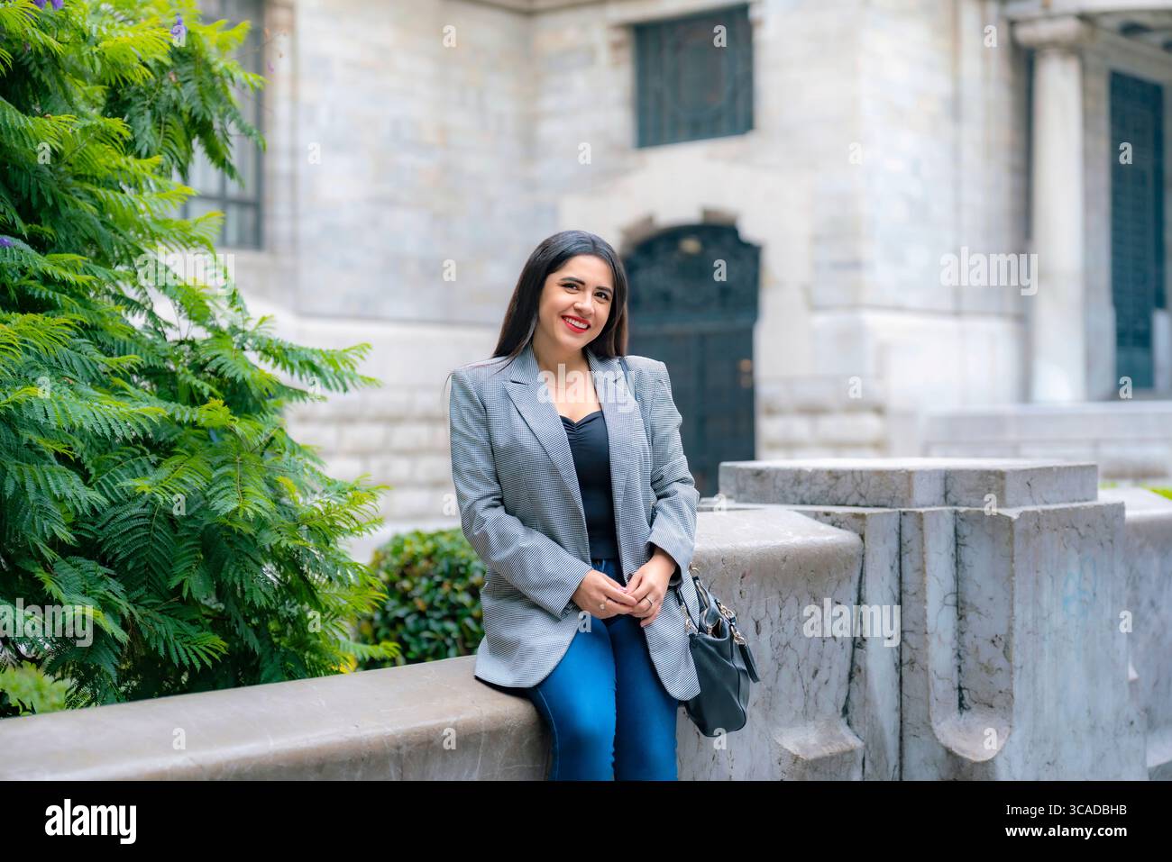Allegra giovane e felice donna d'affari latina che indossa un blazer grigio e pantaloni blu, posa sicura mentre siede vicino alla vecchia architettura del Palacio de Be Foto Stock