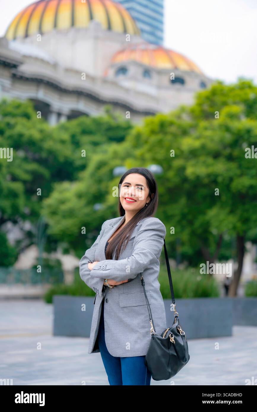 Giovane donna d'affari latina sorridente e felice che posa con le braccia incrociate di fronte al Palacio de Bellas Artes a città del Messico, con un blazer grigio e una pa blu Foto Stock