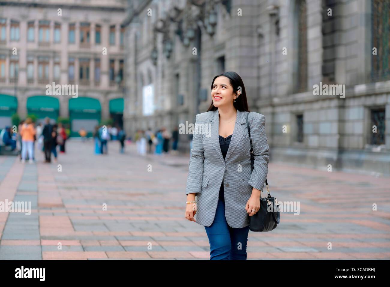 Giovane donna d'affari latina sorridente e felice, con blazer grigio e pantaloni blu, che cammina tranquillamente fuori DAL COMUNE, il Museo Nazionale d'Arte di città del Messico con stile storico Foto Stock