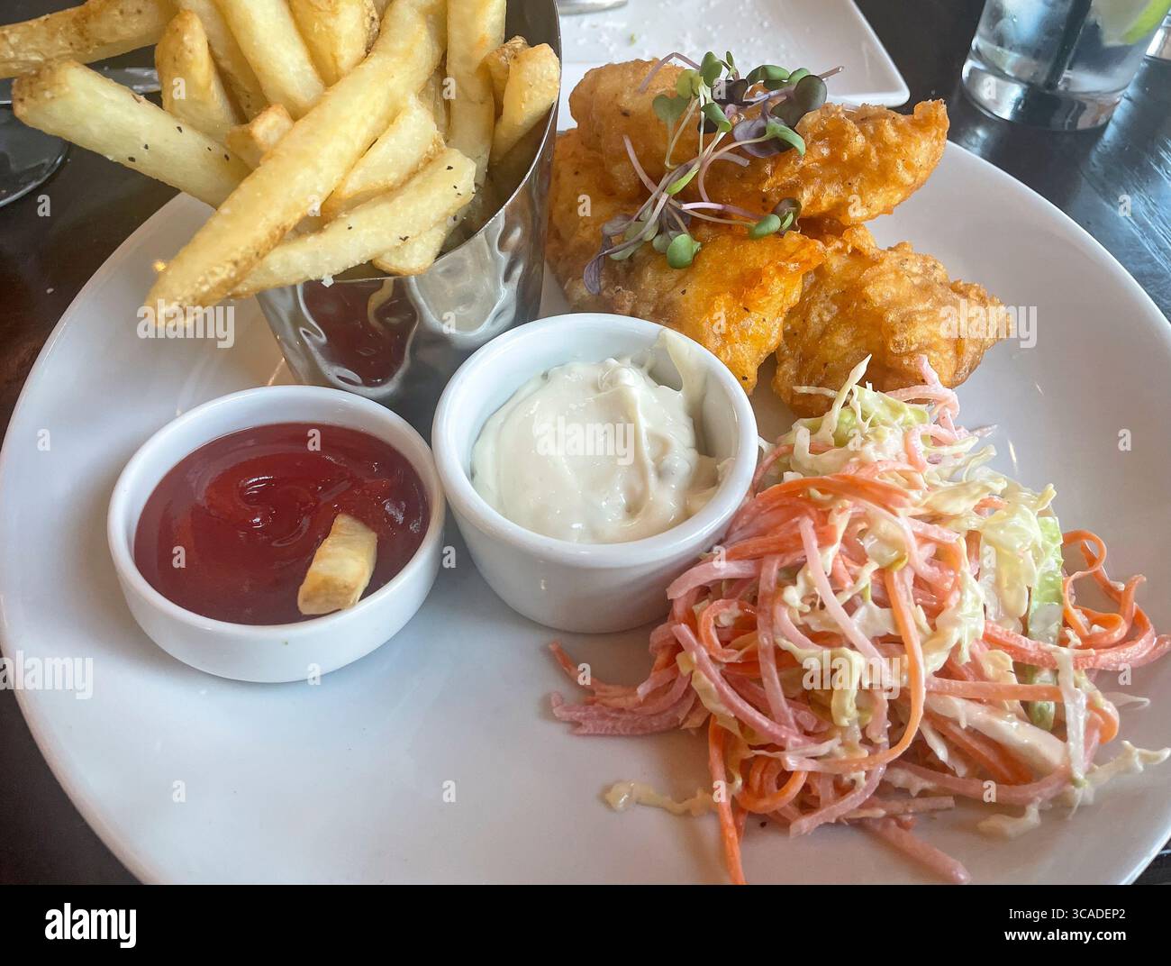 Un classico piatto di fish and chips con filetti di pesce fritti dorati, una tazza di patate fritte in metallo, accompagnato da piccoli camamini bianchi di ketchup e t Foto Stock