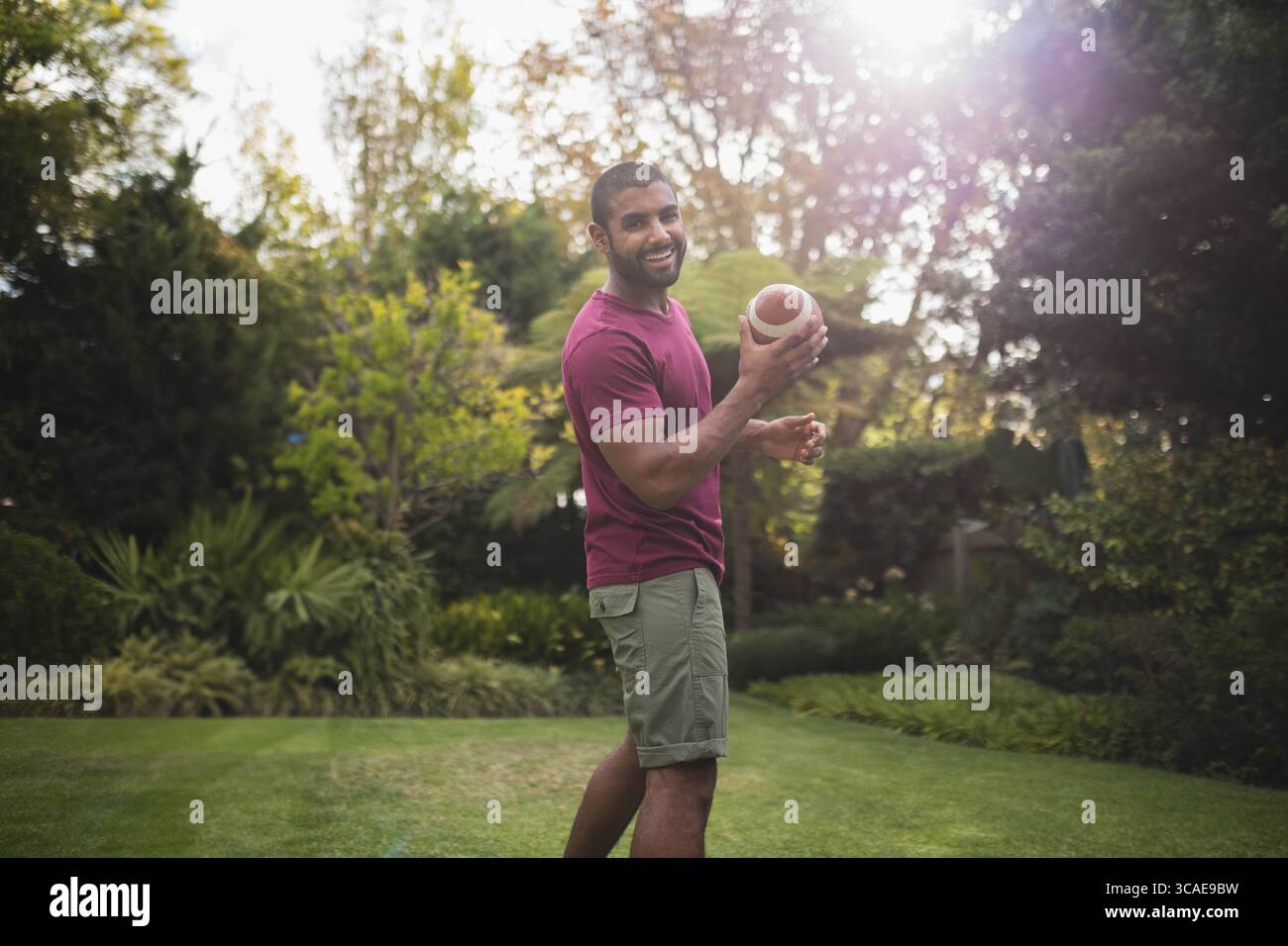 Uomo afro-americano sorridente e tiene il football sul prato del cortile sotto la luce del sole Foto Stock