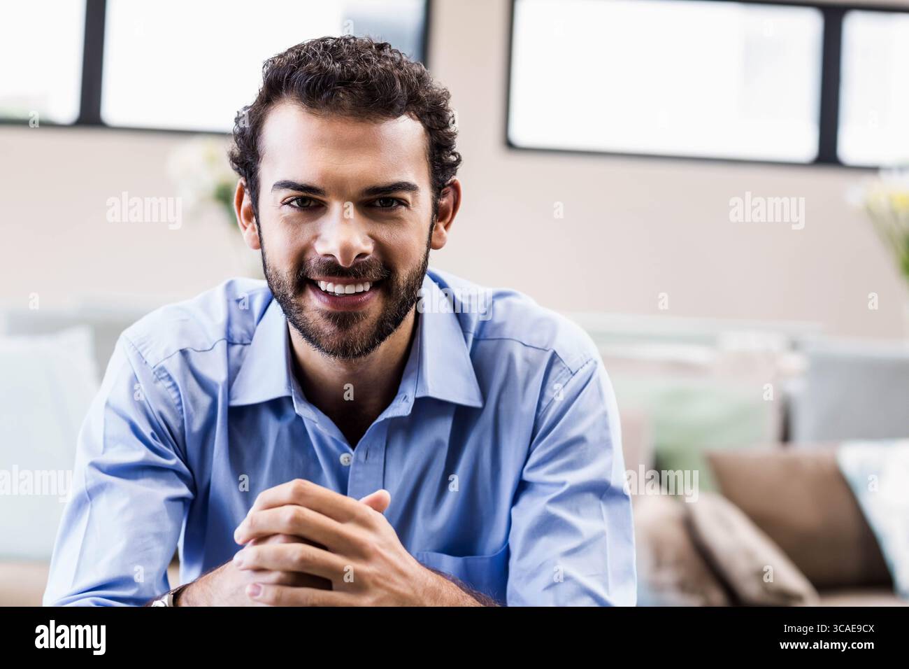 Un uomo d'affari con una camicia in abito sorridente e appoggiato in avanti alla scrivania nel salotto con divani cuscini Foto Stock
