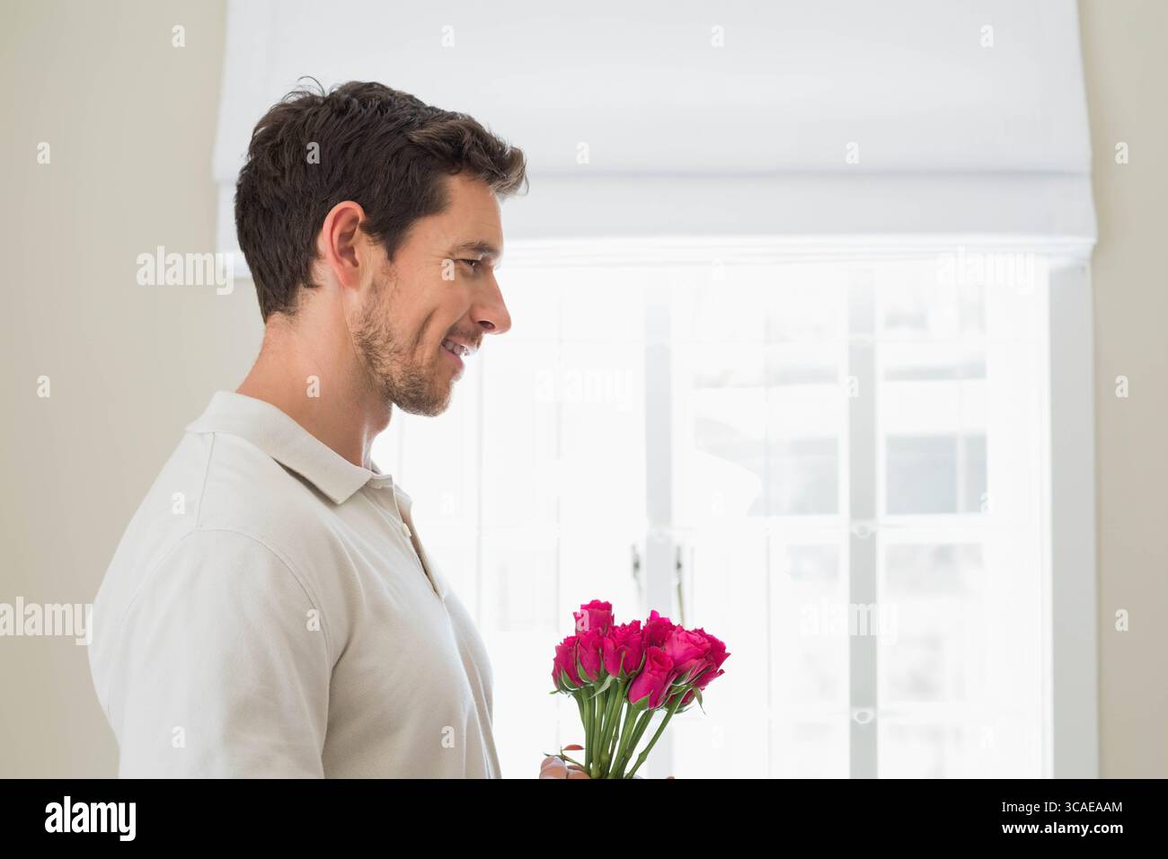 Uomo in piedi in una casa con luce soffusa e con un vivace bouquet di rose rosa vicino alla finestra, copia spazio Foto Stock