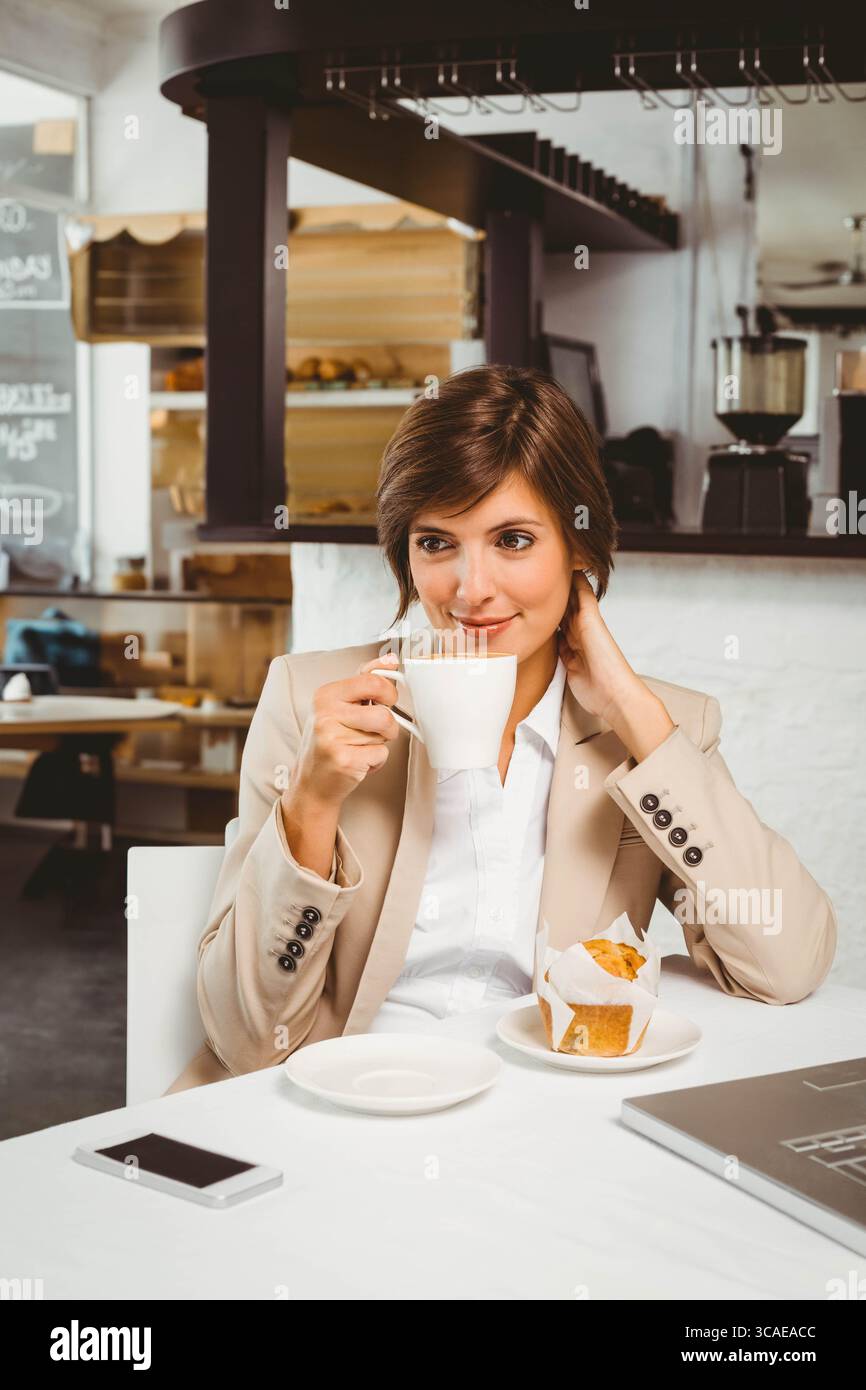 Donna d'affari che indossa un blazer beige che tiene in mano una tazza di caffè mentre srotola il muffin al tavolo del caffè Foto Stock