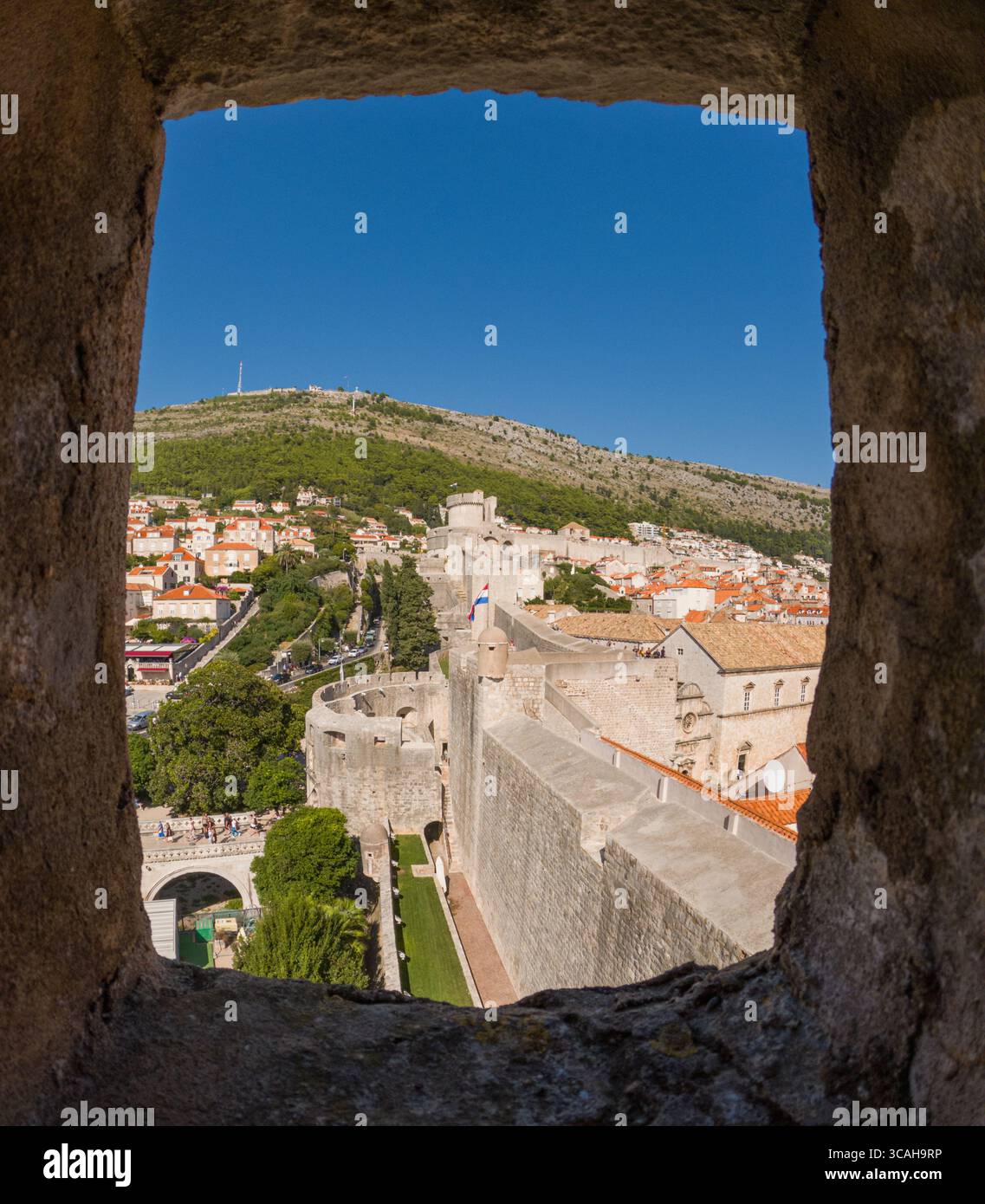 Dubrovnik, Croazia - 14 settembre 2023: Una vista delle mura della città e del Monte Srd attraverso una finestra di pietra. I turisti camminano lungo le mura, esplorando il Foto Stock