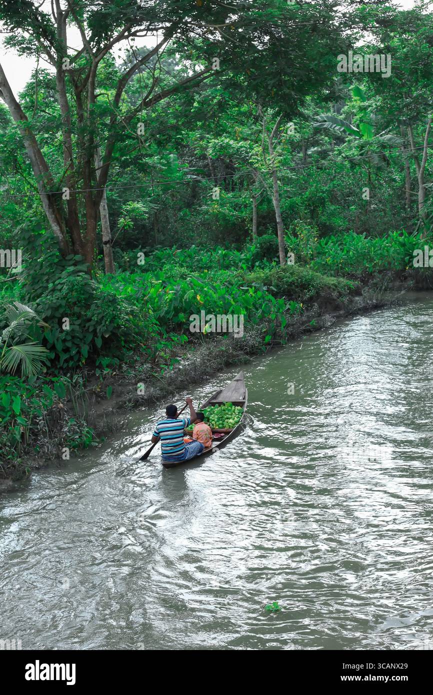 Barishal, Bangladesh - 09 agosto 2018: Vista di una barca solitaria che attraversa il fiume grigio, incorniciato dal lussureggiante e vibrante fogliame verde della riva del fiume. Foto Stock