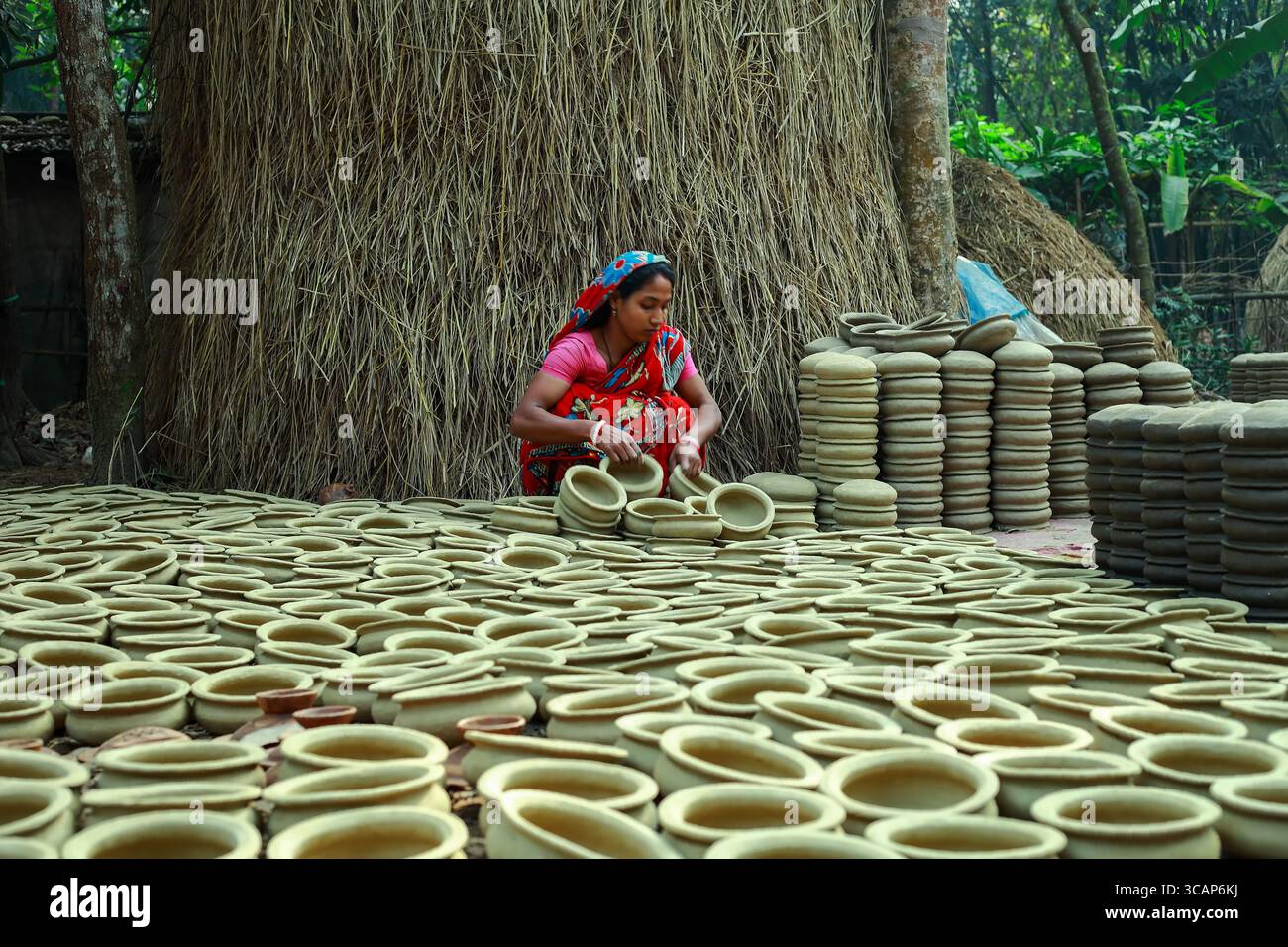 Cumilla, Bangladesh - 25 gennaio 2020: Veduta di una donna in un abbigliamento vivace in mezzo a un mare di vasi di terra, le sue mani modellano l'argilla sotto il sole caldo, sullo sfondo di rustici fienili. Foto Stock