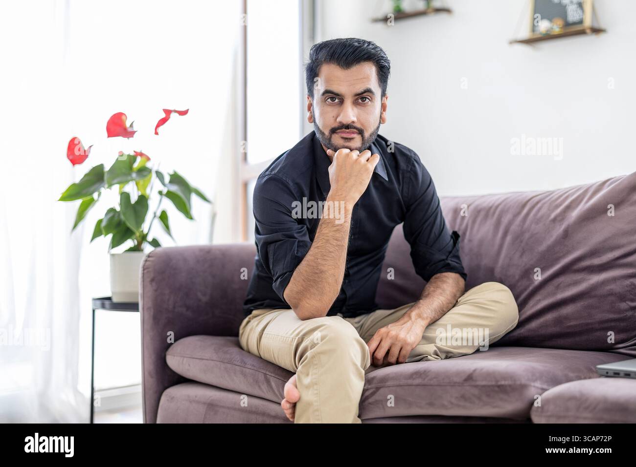 Uomo barbuto seduto su un divano nel salotto di casa sua e guardando la macchina fotografica Foto Stock
