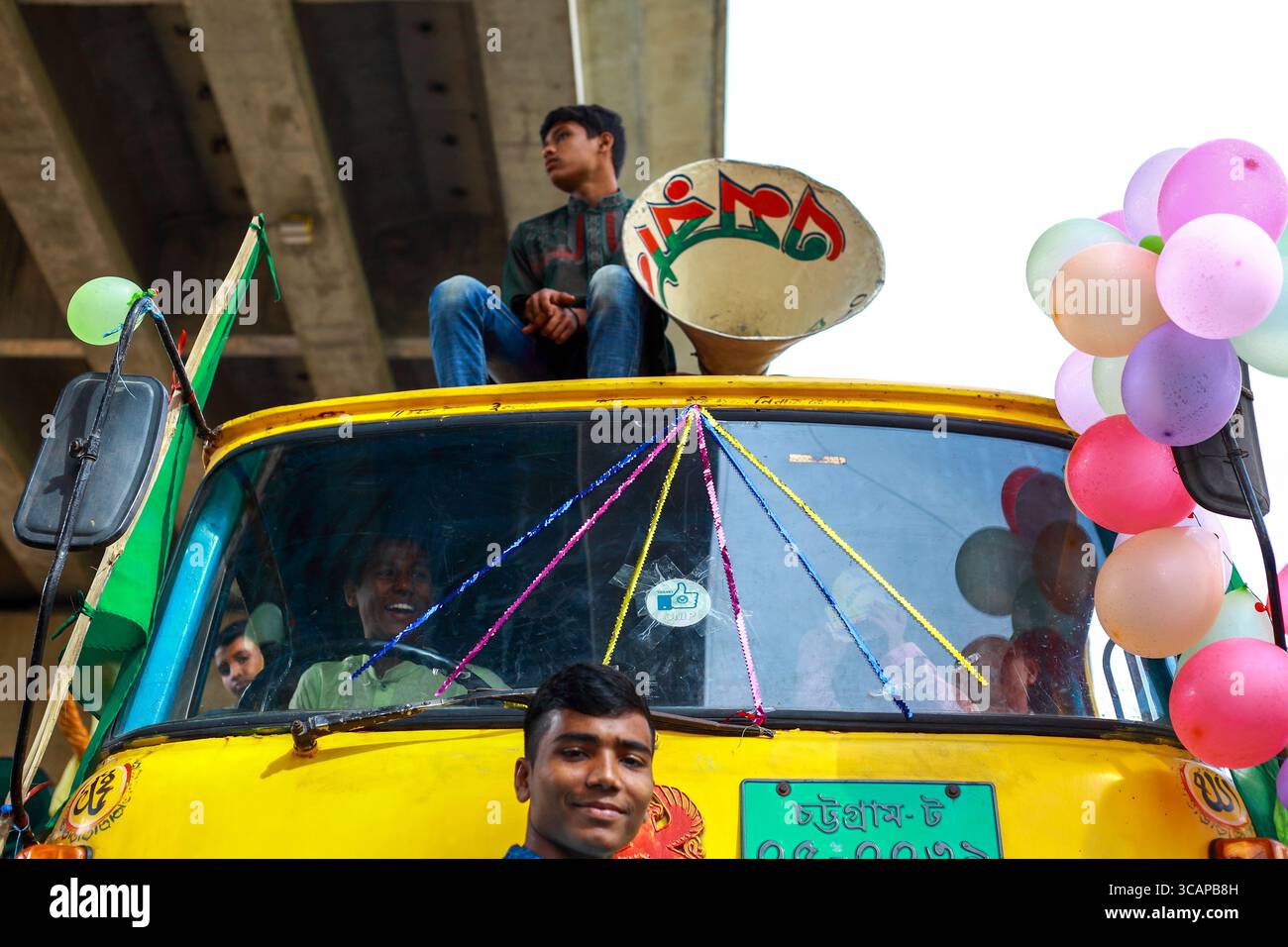 Chattogram, Bangladesh - 21 novembre 2018: Vista di un lucente risciò giallo CNG adornato da palloncini colorati e un giovane arroccato in cima, sotto l'ombra fresca di un cavalcavia di cemento. Foto Stock