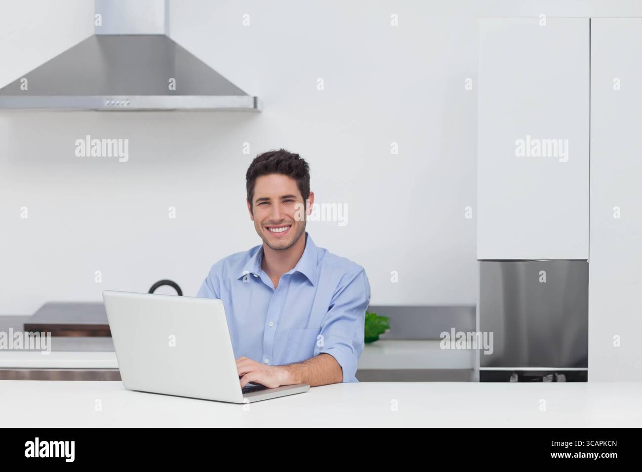 Uomo seduto su un piano di lavoro bianco in una cucina moderna che lavora su un computer portatile argento vicino a una pianta verde Foto Stock