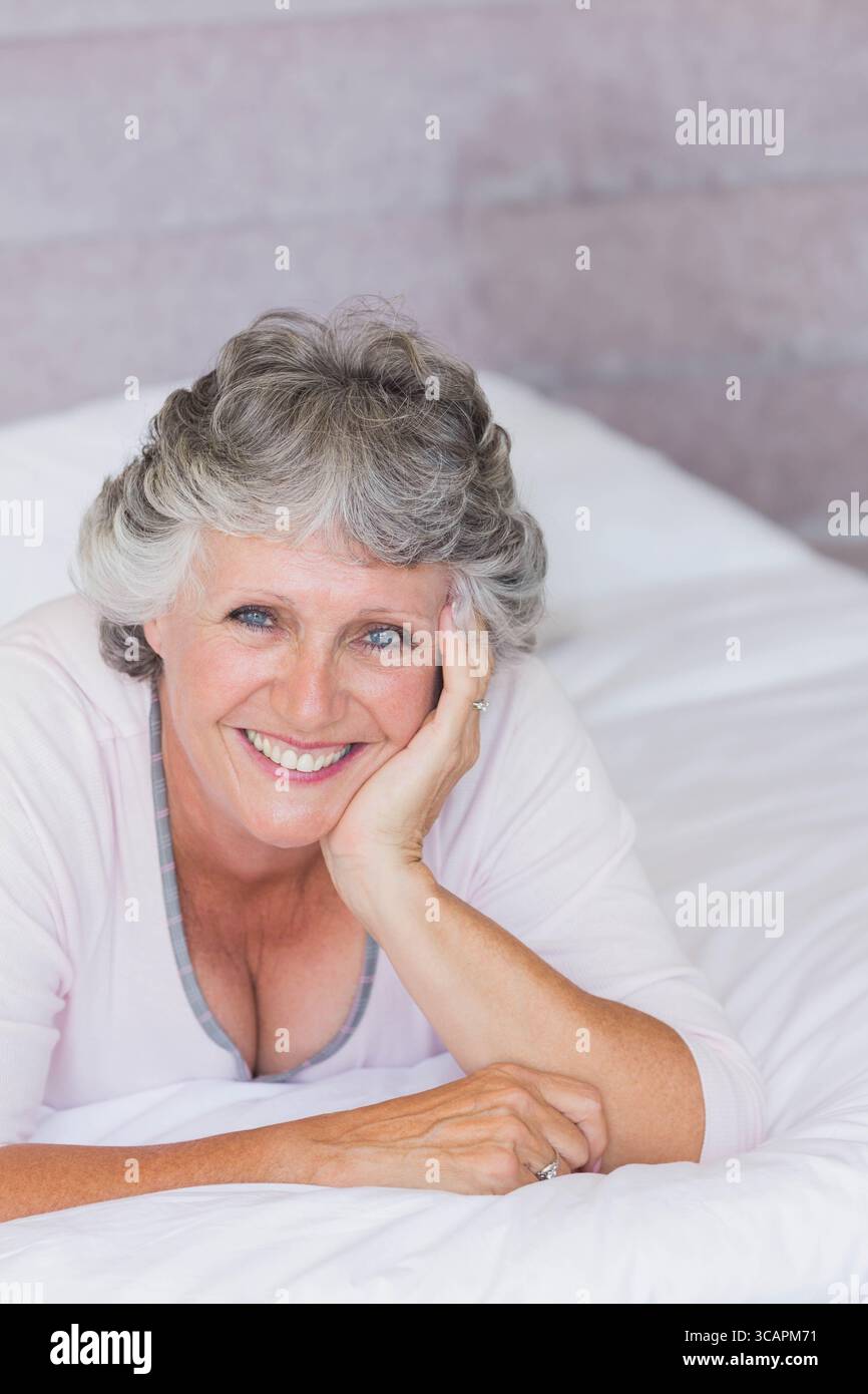 Donna anziana sorridente mentre si sdraia sul letto con lenzuola bianche in camera da letto che mostrano un anello a portata di mano Foto Stock