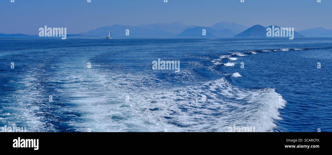 La barca si sveglia in un bellissimo blu profondo con uno sfondo delle montagne di Cefalonia e una barca a vela Foto Stock