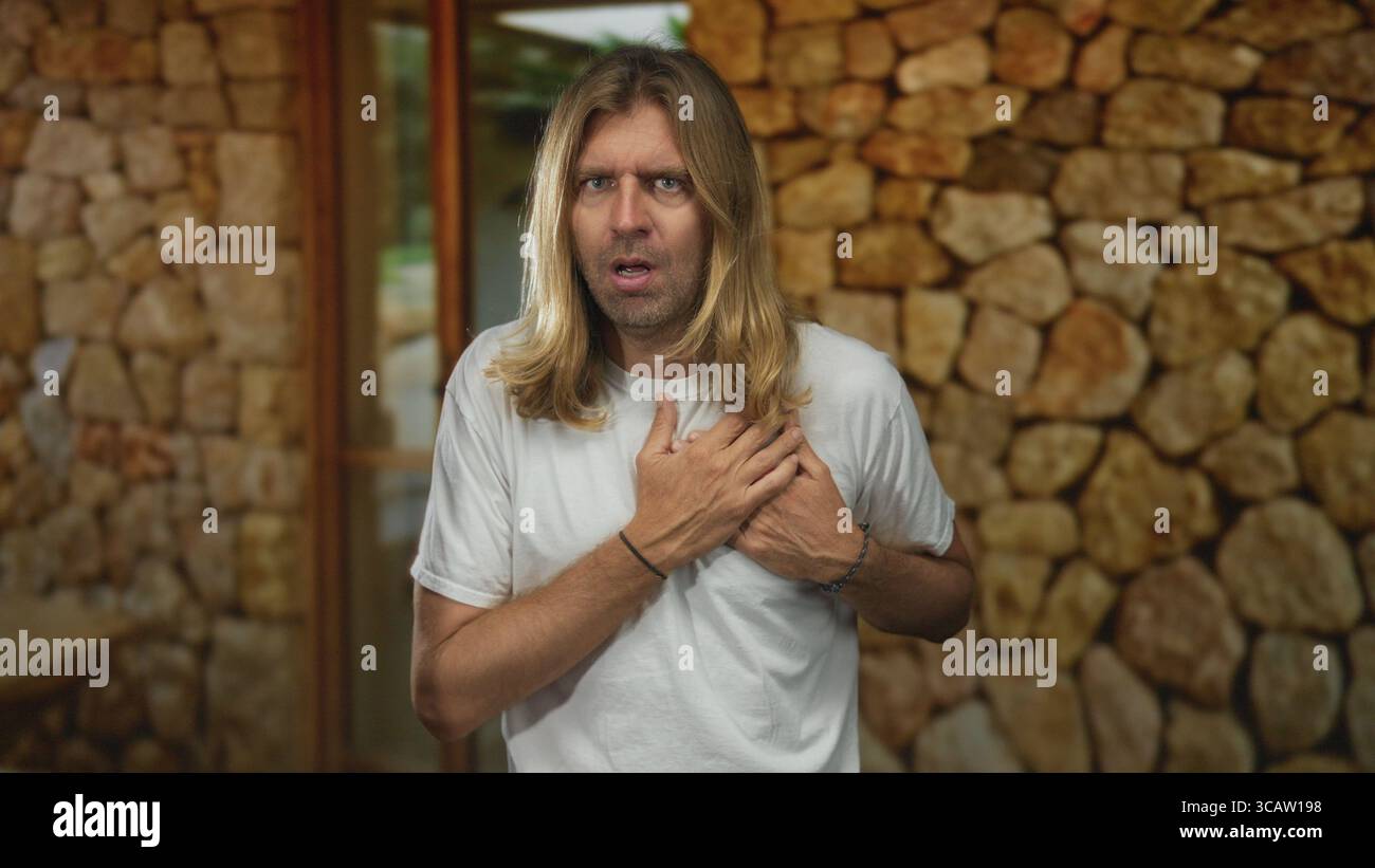L'uomo biondo con i capelli lunghi stringe il petto per attacco di cuore mentre è in piedi alla porta di ingresso dell'edificio incorniciato da un muro di pietra; attacco di cuore vulnerabilità di soccorso Foto Stock