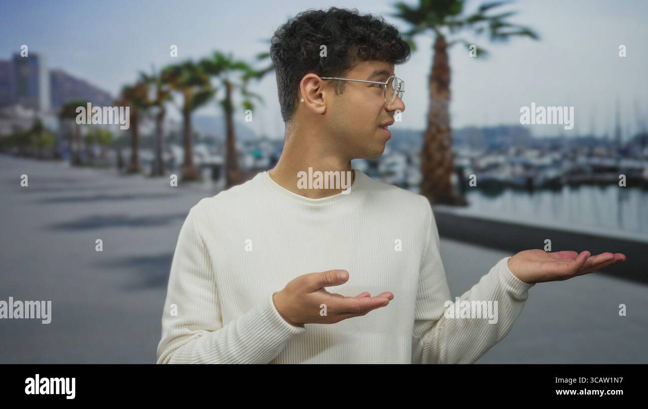 L'uomo che indossa un maglione bianco e occhiali mostra le palme rivolte verso l'alto accanto alla barca sul lungomare sotto le palme; incertezza. Foto Stock