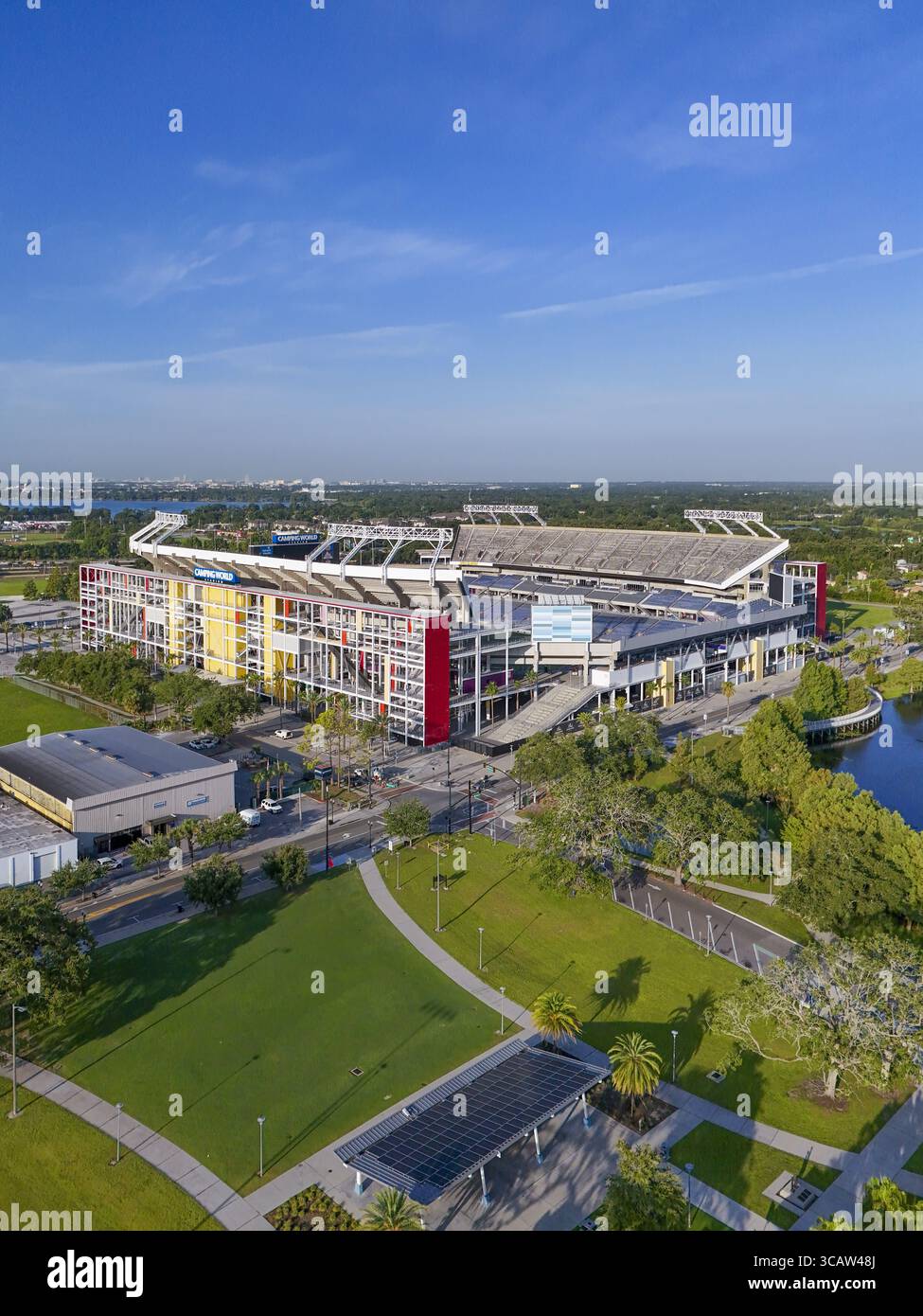 Orlando, Stati Uniti - 21 luglio 2025: Veduta aerea del Camping World Stadium crogiolandosi sotto un cielo limpido, con i suoi vibranti accenti rossi e gialli che si stagliano contro il lussureggiante paesaggio verde. Foto Stock