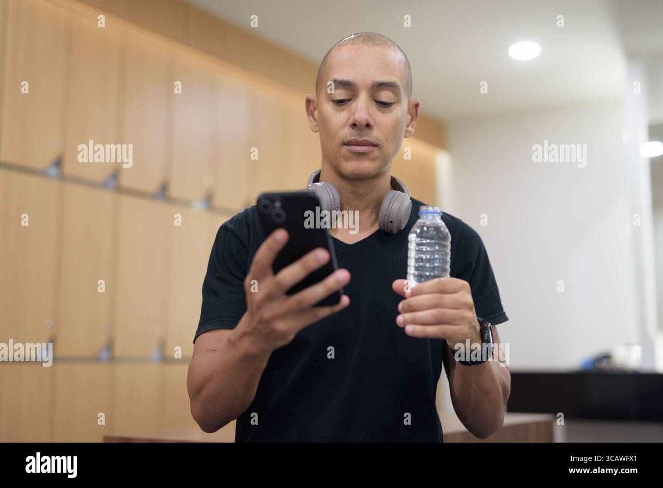 Uomo latino adulto in spogliatoio che tiene una bottiglia d'acqua mentre utilizza lo smartphone. Recupero dell'allenamento in palestra, abitudine all'idratazione, routine di fitness digitale, privacy A. Foto Stock