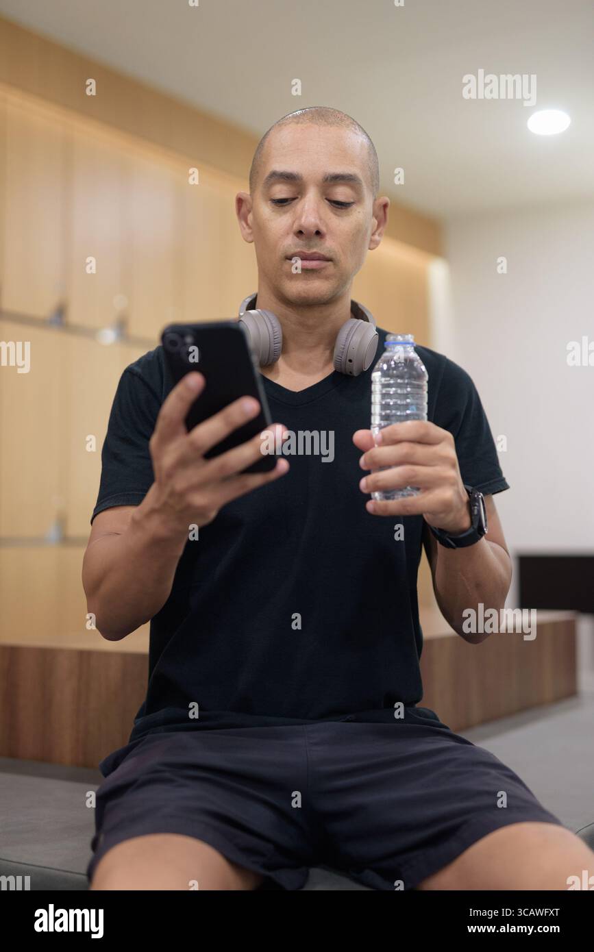 Uomo latino adulto in spogliatoio che tiene una bottiglia d'acqua mentre utilizza lo smartphone. Recupero dell'allenamento in palestra, abitudine all'idratazione, routine di fitness digitale, privacy A. Foto Stock