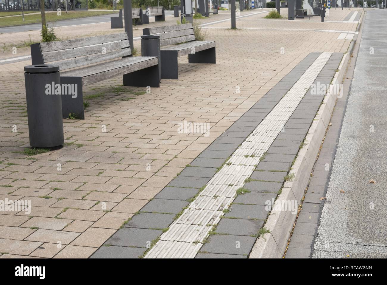 La pavimentazione tattile guida i pedoni alla fermata dell'autobus dotata di panchine moderne e lattine per la spazzatura Foto Stock