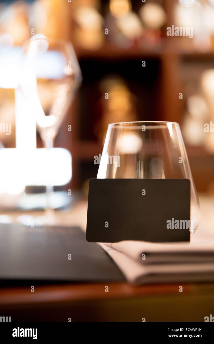 Un elegante biglietto da visita nero si erge accanto a un vetro, con uno sfondo sfocato del ristorante che crea un'atmosfera sofisticata. Questo mockup presenta un p Foto Stock