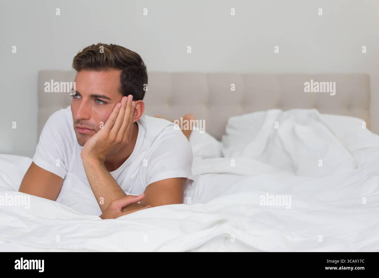 Uomo ispanico sdraiato sul letto con biancheria da letto bianca in camera da letto con maglietta bianca vicino alla testata del letto trapuntata Foto Stock