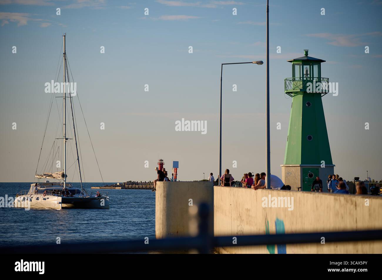 Barca a vela che lascia il porto vicino al faro verde con gente che guarda al tramonto a Danzica. Foto Stock