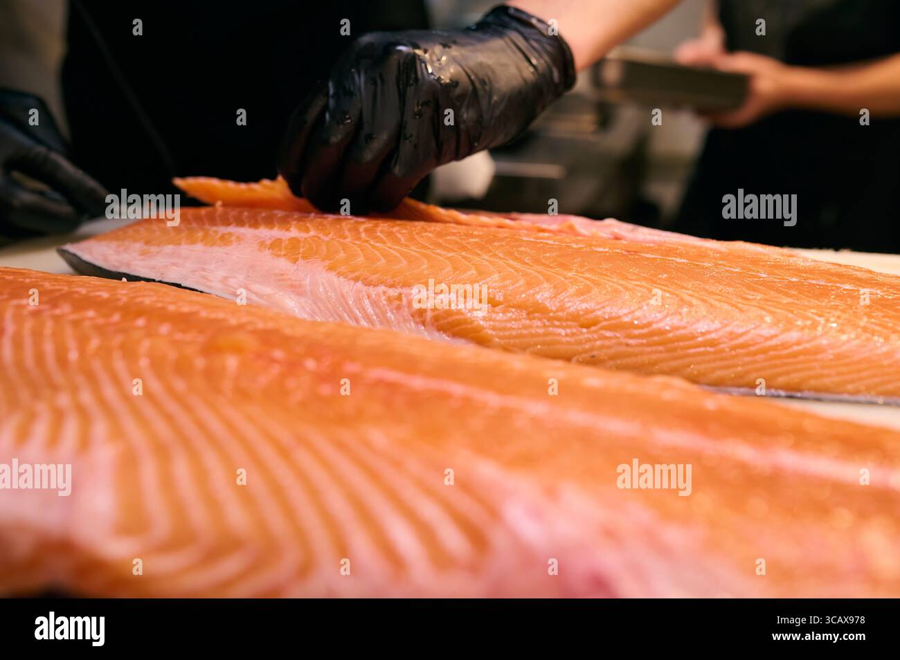 Lo chef affetta con precisione il filetto di salmone fresco su un tagliere in una cucina professionale. Primo piano delle mani con guanti neri e consistenza di pesce crudo. Foto Stock