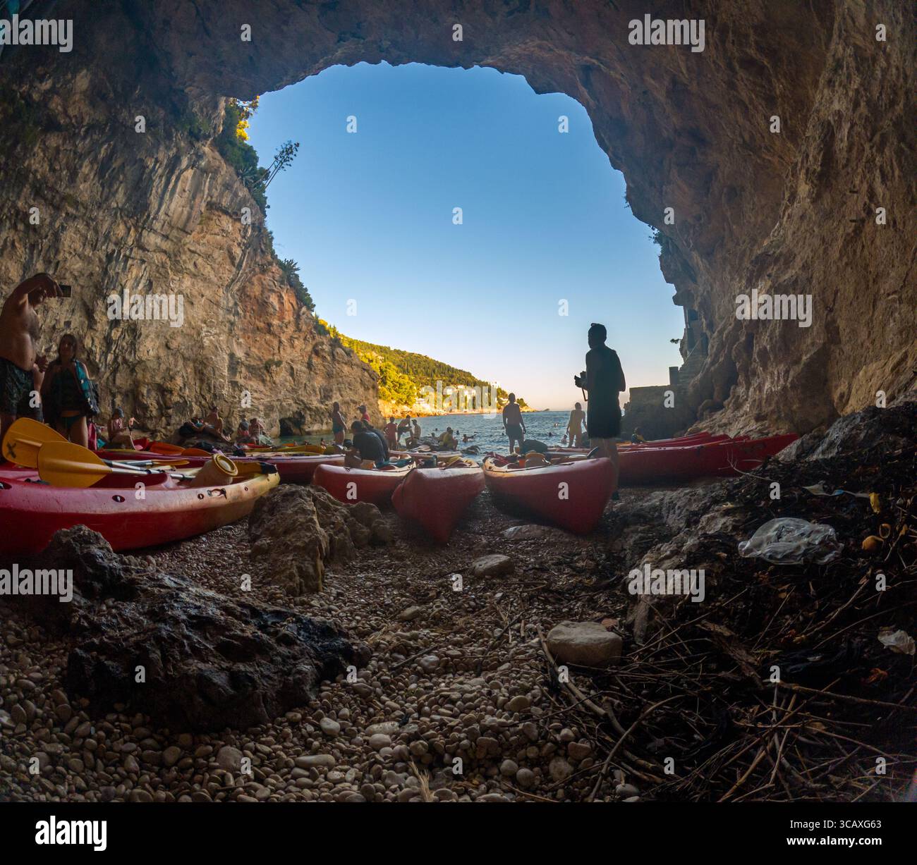 Dubrovnik, Croazia - 12 settembre 2023: I kayak si riuniscono all'interno di una grotta durante un tour in kayak al tramonto. Pagaie e kayak sono visibili sulla spiaggia rocciosa. Foto Stock