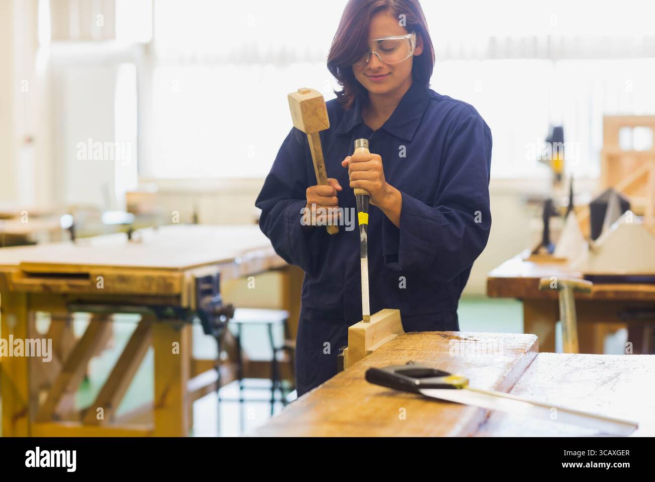 Donna asiatica che indossa occhiali di protezione e tuta scalpellare blocco di legno con mazzuolo, scalpello alla panca del negozio di legno Foto Stock