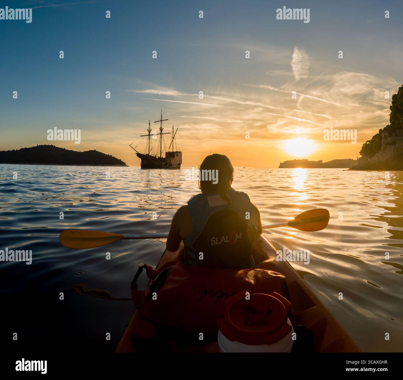 Dubrovnik, Croazia - 12 settembre 2023: Un kayaker gode di un tour al tramonto, pagaiando davanti a una replica di galeoni vicino alla costa della città. Foto Stock