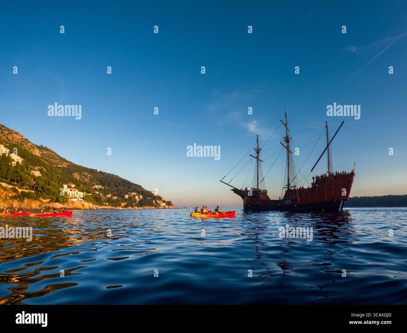 Dubrovnik, Croazia - 12 settembre 2023: I kayak pagaiano vicino a una replica di galeone durante un tour al tramonto, godendosi le viste panoramiche e le acque calme. Foto Stock