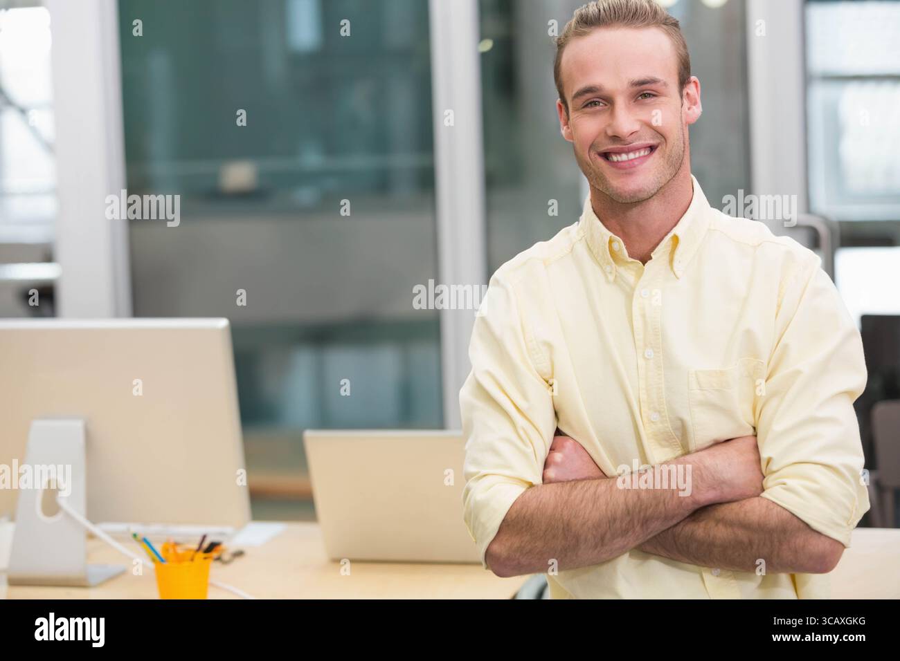 Dipendente maschio in piedi dietro una scrivania in legno in un ufficio moderno con monitor, laptop e porta penna arancione Foto Stock
