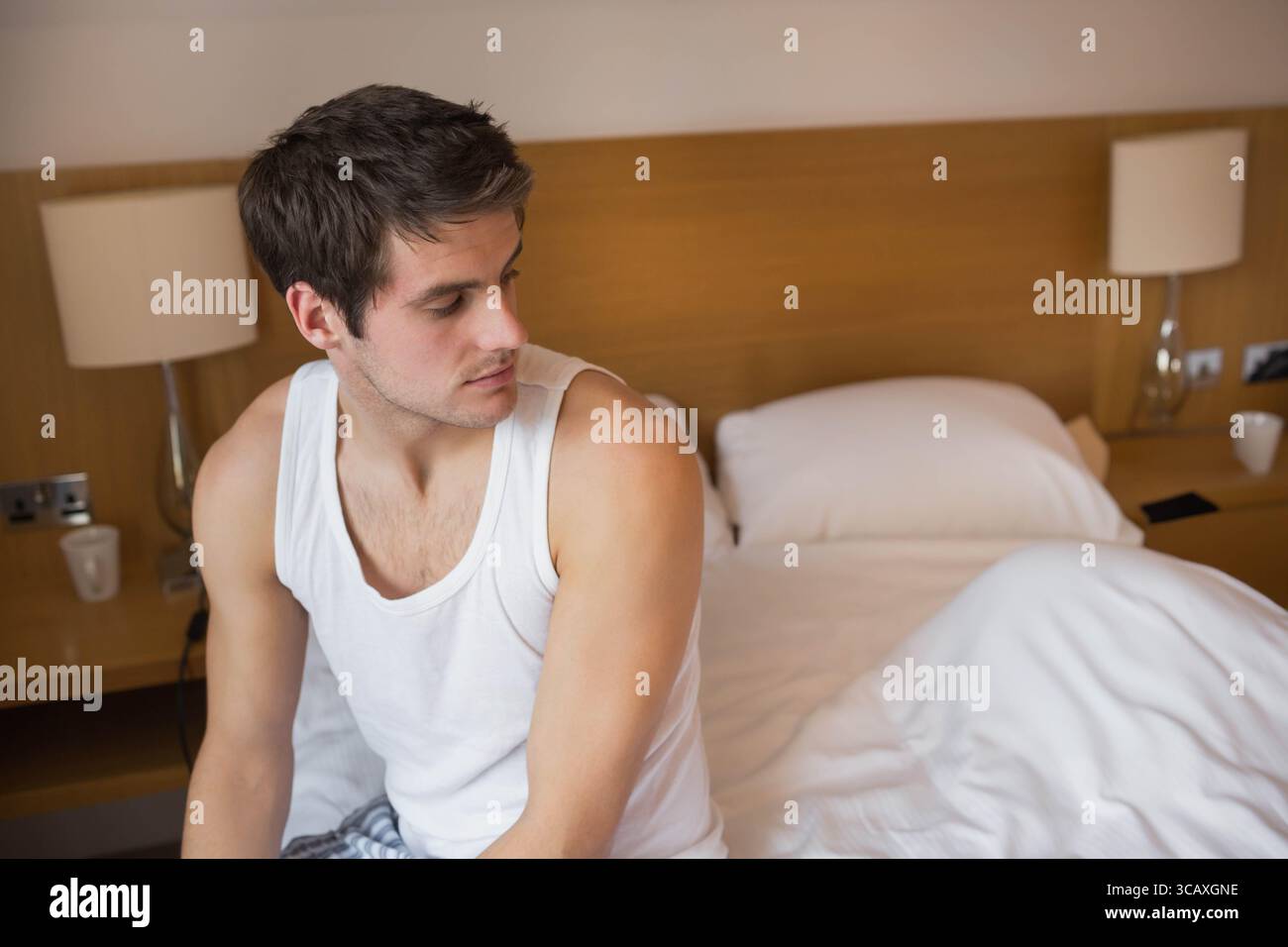 Uomo seduto sul bordo di un letto non fatto in una camera da letto in stile hotel con pigiama che controlla lo smartphone Foto Stock