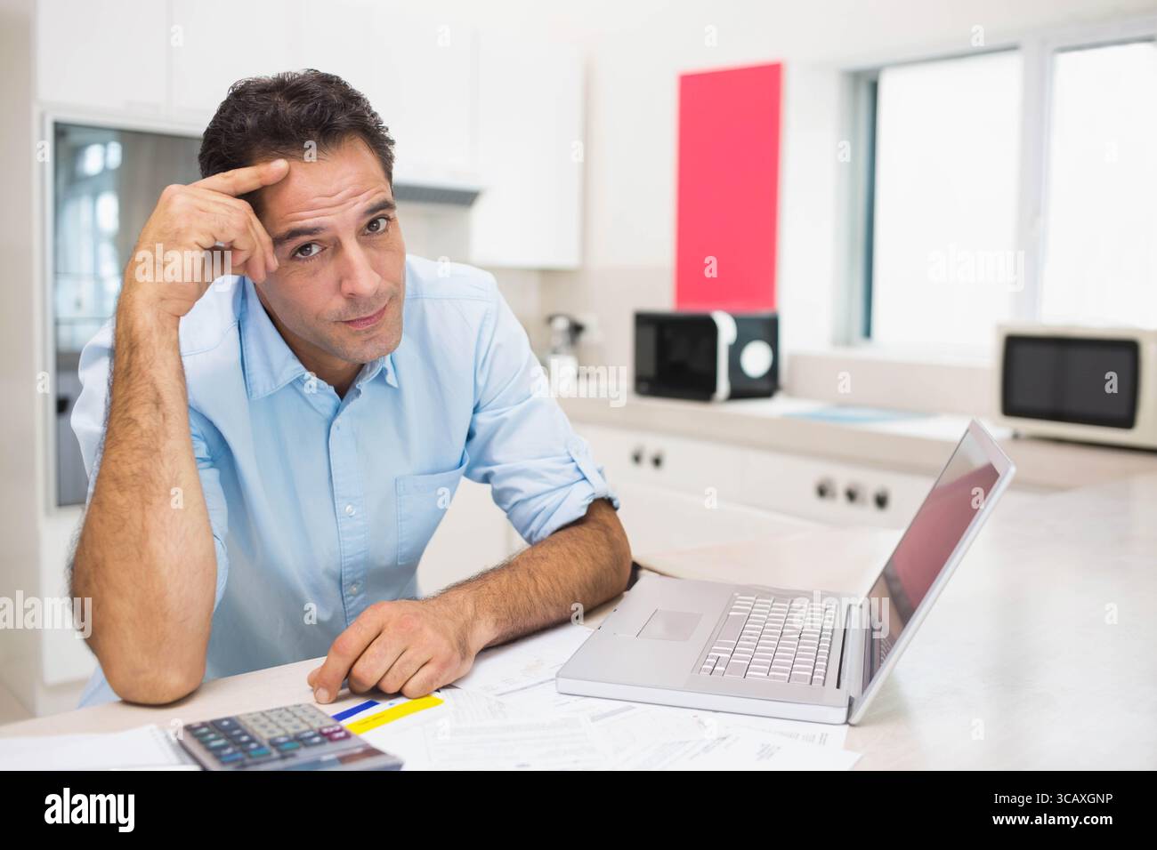 Uomo di mezza età seduto nella moderna isola della cucina a rivedere le bollette con calcolatrice, penna e laptop Foto Stock
