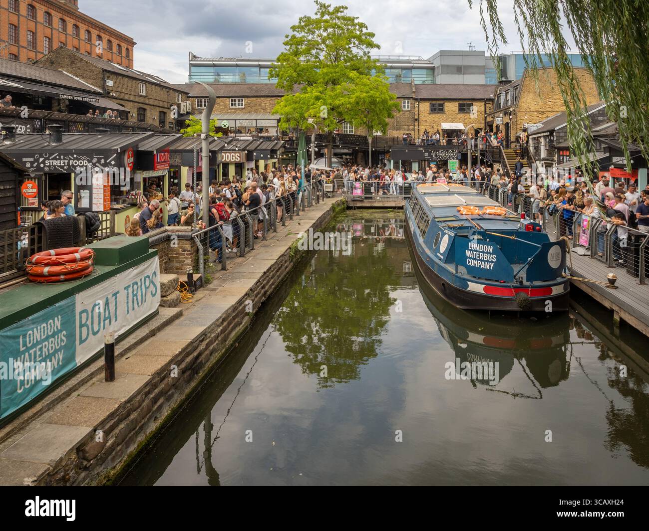 Una barca sul canale della London Waterbus Company è ormeggiata sul Regent's Canal tra le vivaci bancarelle di Street food di Camden Market, Londra, Regno Unito. Foto Stock