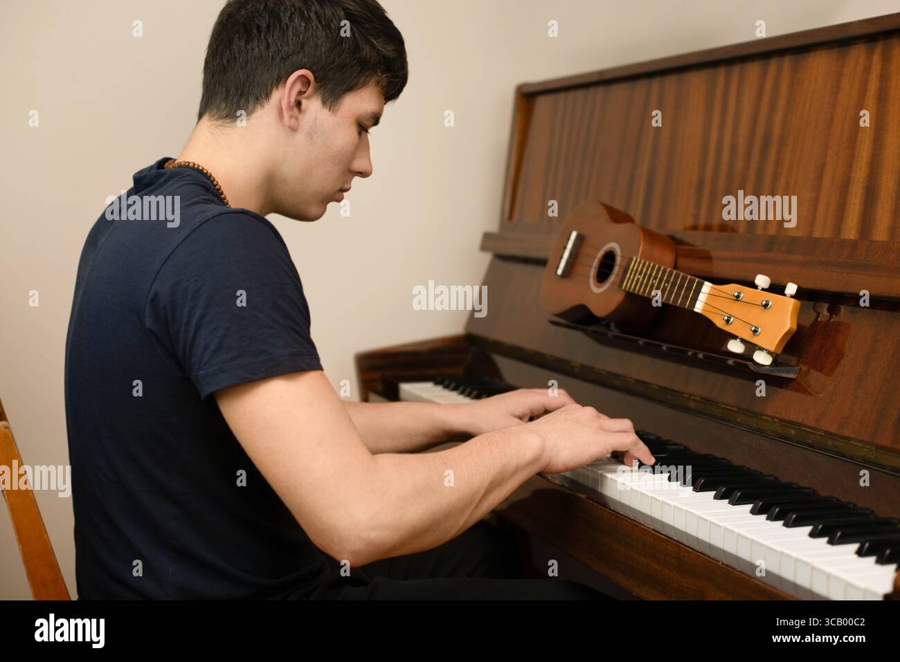 Ragazzo seduto da un pianoforte che si concentra sulla musica, con un ukulele sopra lo strumento. Questa immagine rappresenta la dedizione all'apprendimento Foto Stock