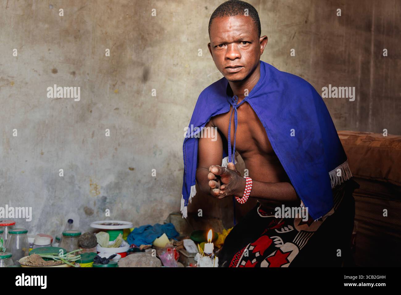 Sangoma del villaggio africano che indossa un mantello blu e un abbigliamento tradizionale, eseguendo un rituale spirituale all'interno, circondato da erbe, ossa e candele, B. Foto Stock