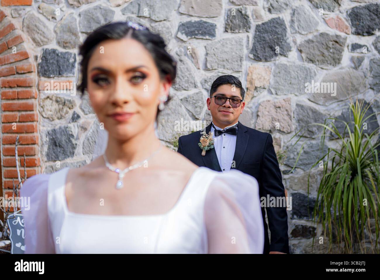 Su un muro rustico in pietra, la sposa sfocata in primo piano e lo sposo concentrato sullo sfondo creano un ritratto artistico ed elegante. Foto Stock