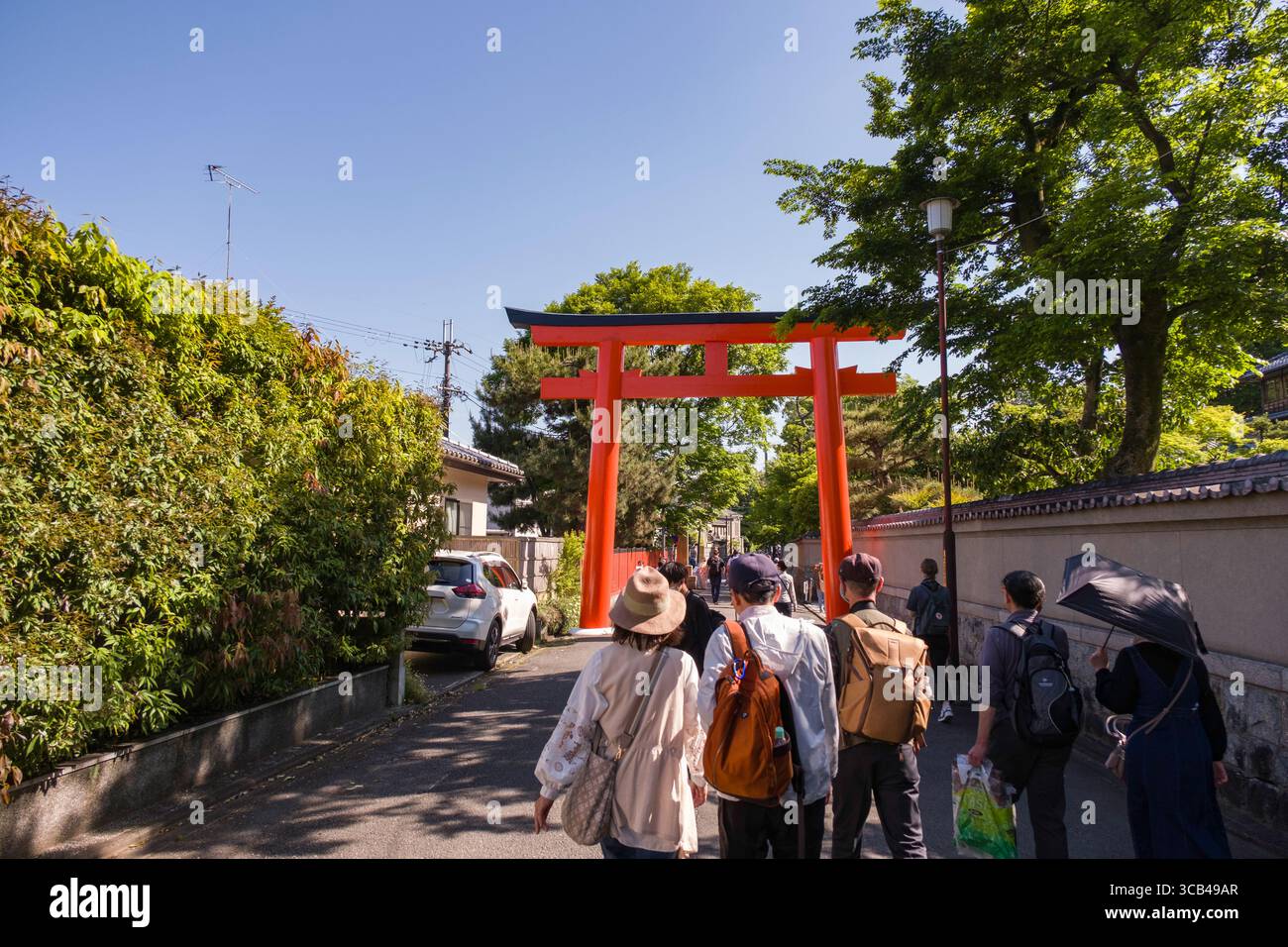 Un gruppo di turisti cammina attraverso una strada giapponese, avvicinandosi a una vivace porta torii rossa, circondata da vegetazione lussureggiante, in una giornata di sole Foto Stock