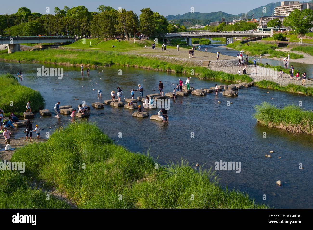 Persone che trascorrono una giornata di sole usando pietre per attraversare il fiume Kamo sul delta di Kamogawa. La vegetazione lussureggiante e l'acqua che scorre creano un tranquillo e. Foto Stock