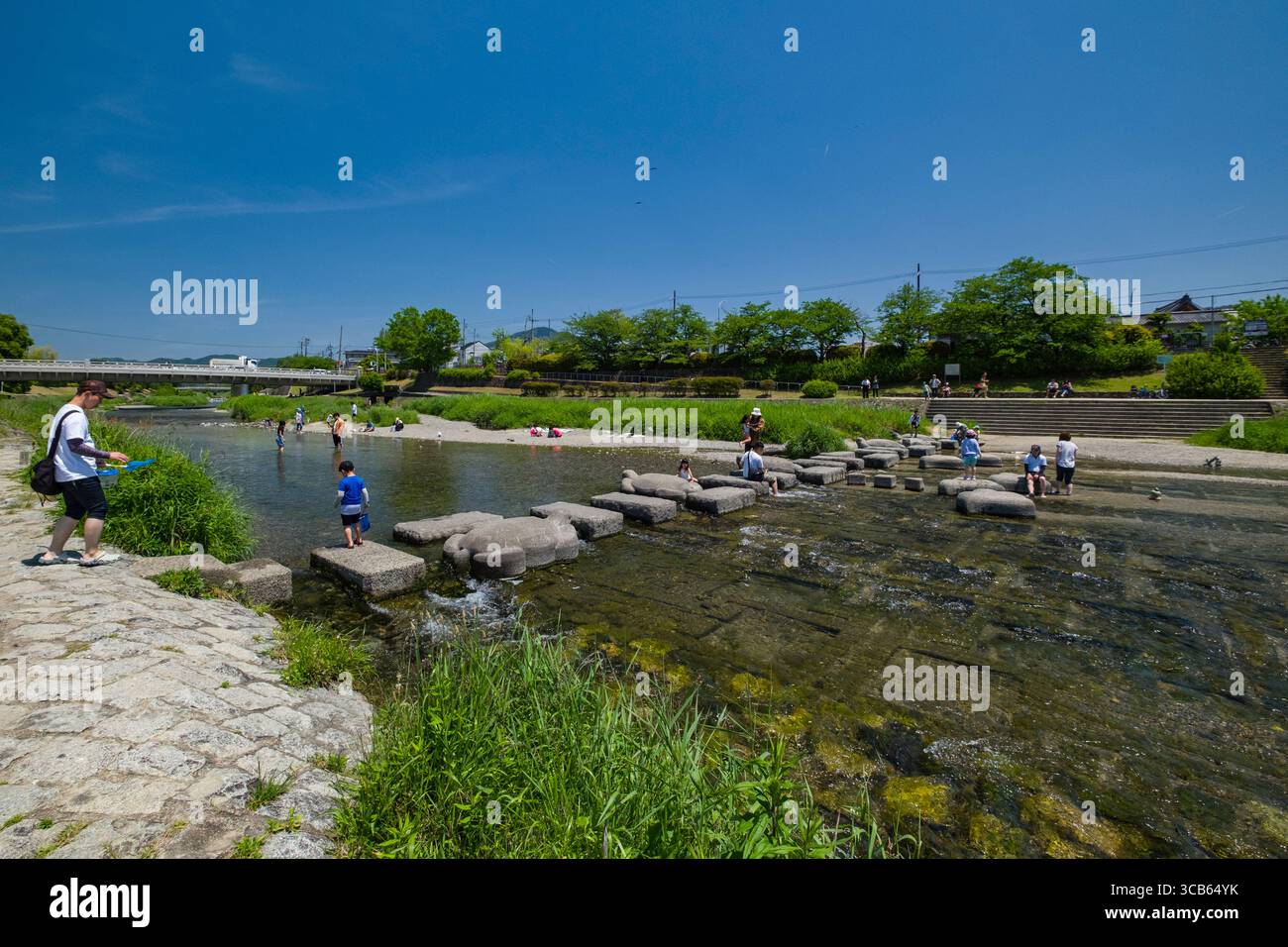 Un ambiente tranquillo con famiglie e bambini che esplorano un pittoresco parco lungo il fiume sotto un cielo azzurro. Le persone attraversano pietre di gradini e si godono il re Foto Stock