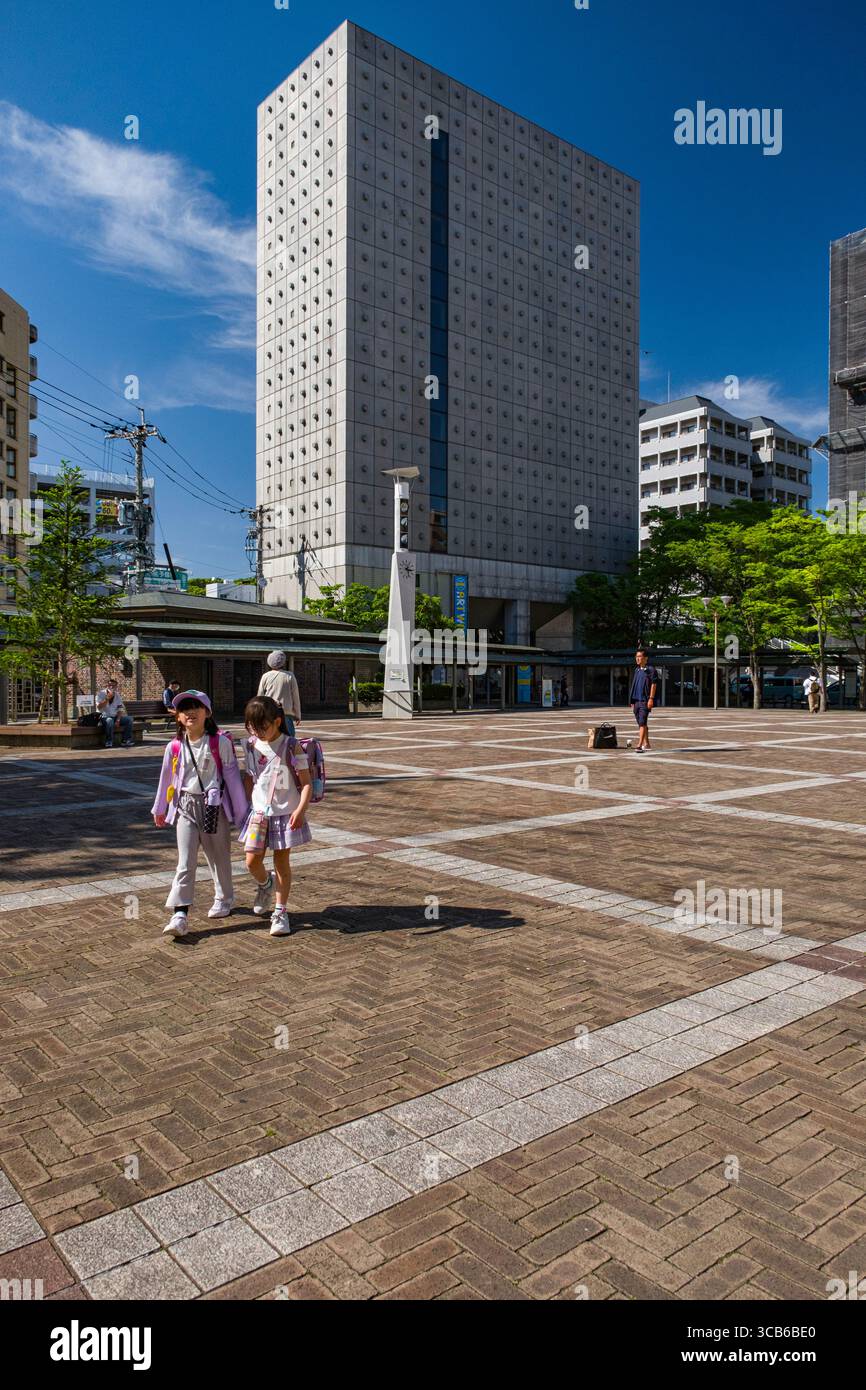 Due bambini con zaini camminano in una piazza soleggiata circondata da edifici moderni, che simboleggiano l'esplorazione e la vita quotidiana, Sasebo, Giappone Foto Stock