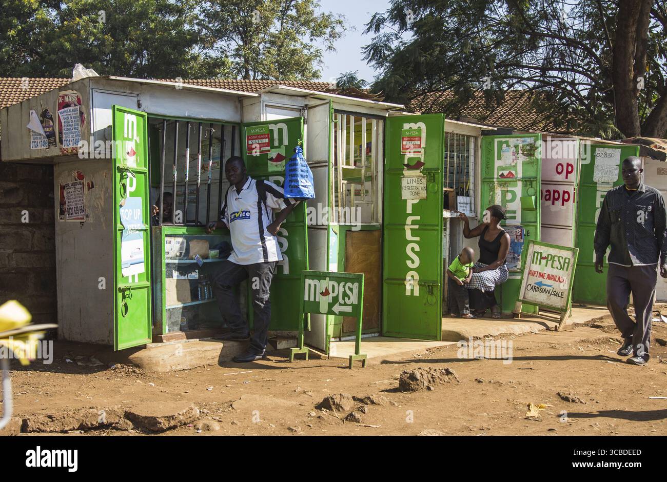 Kisumu, Kenya - 13 maggio 2021: Vista dei chioschi verdi e vibranti M-pesa fiancheggiano una strada polverosa, piena di attività sotto il caldo sole keniota. Foto Stock